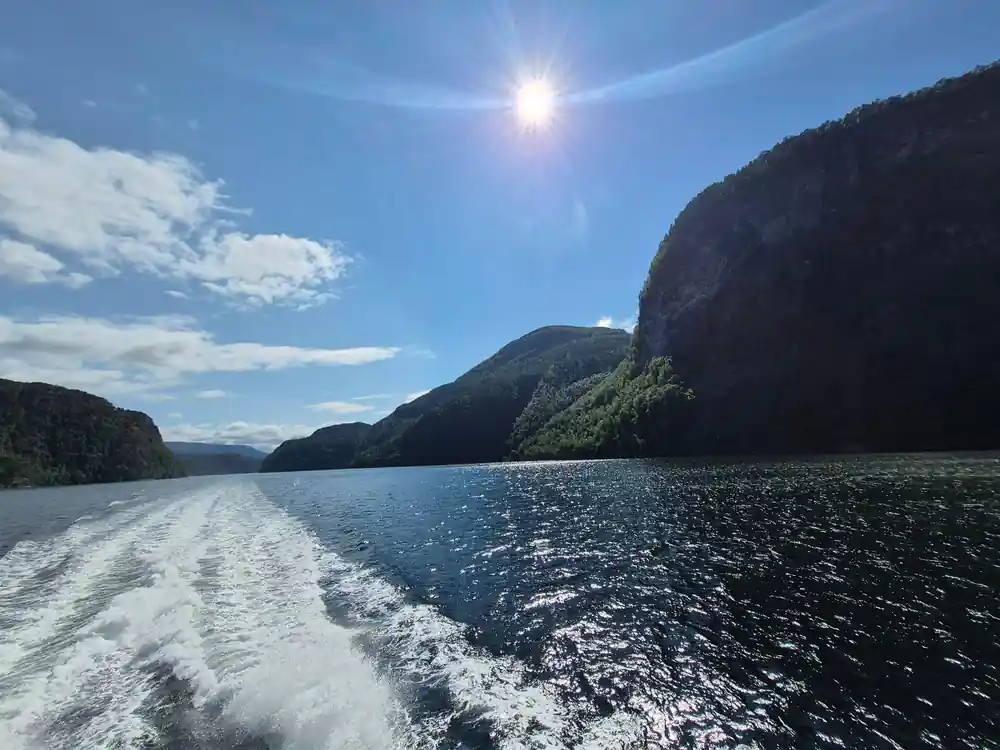 The sun high over the wake as the fjord opens behind